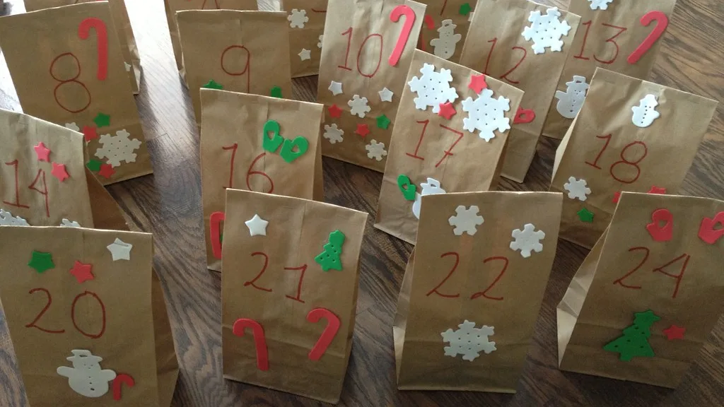 Nummerierte braune Papiertüten mit aufgeklebten Schneeflocken und Sternen stehen auf einem Holzboden, die Zahlen 8 bis 24 sind lesbar