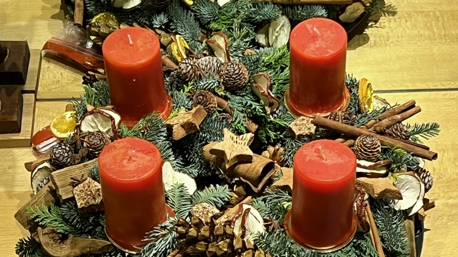 Selbst gebundener Adventskranz aus Tannengrün mit vier roten Kerzen, Zapfen, Zimtstangen und getrockneten Orangenscheiben auf einem hellen Holztisch