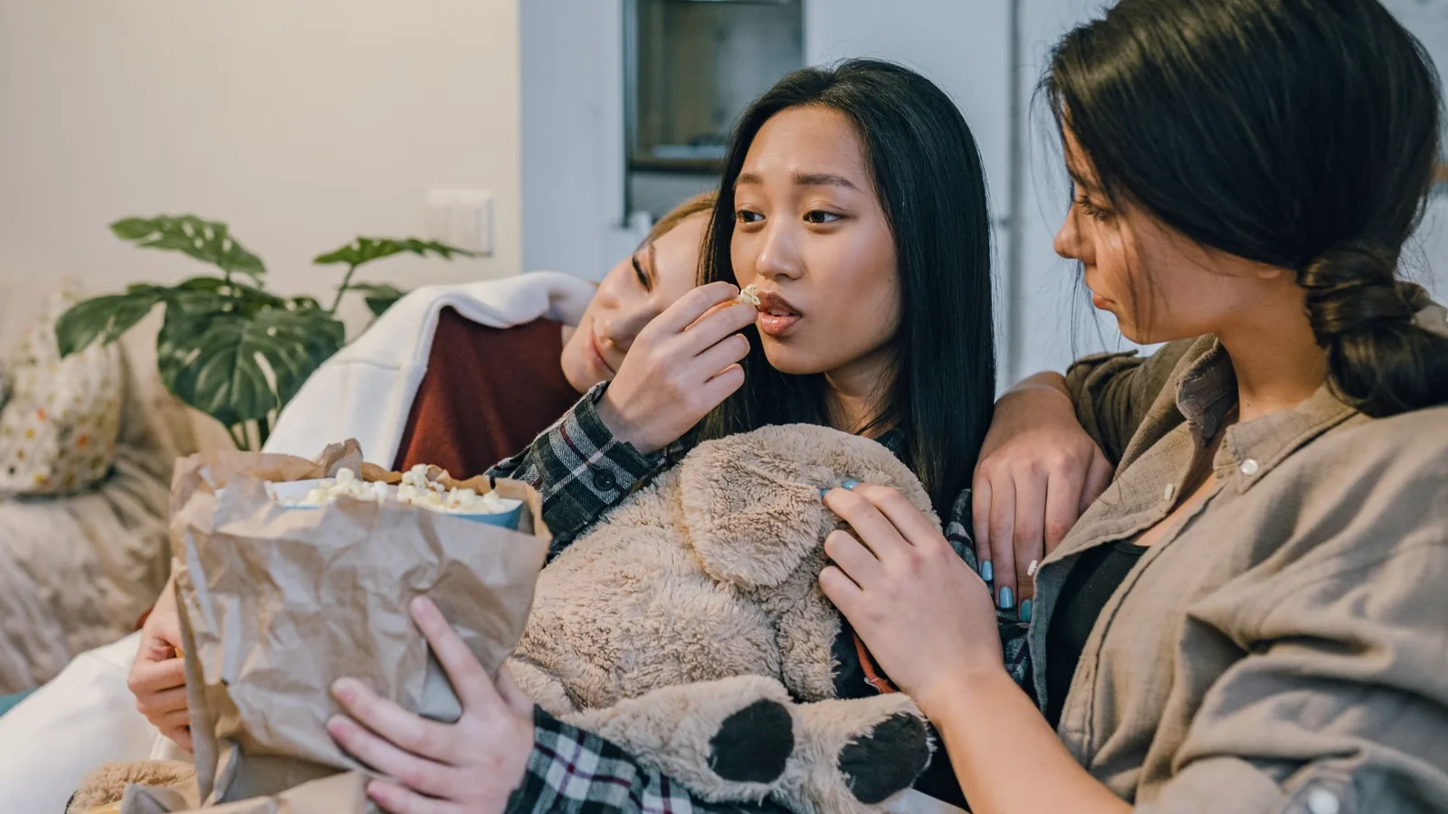 Drei Menschen sitzen mit Popcorn und einer Decke auf einem Sofa, warmes Tageslicht, kein Bildschirminhalt sichtbar