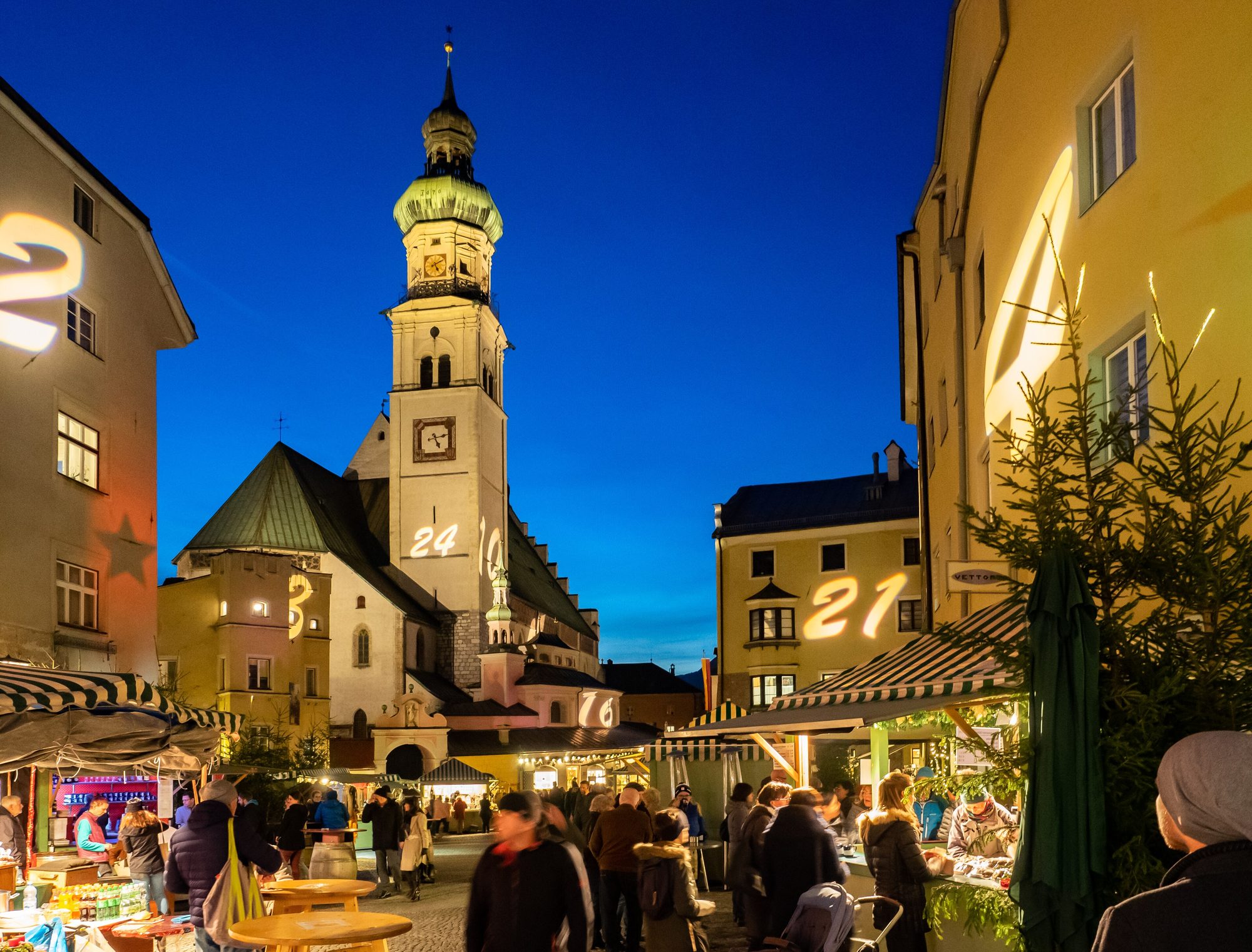 Beleuchtete Stände des Adventmarkts in der Altstadt von Hall in Tirol zur blauen Stunde, dahinter der Turm der Stadtpfarrkirche