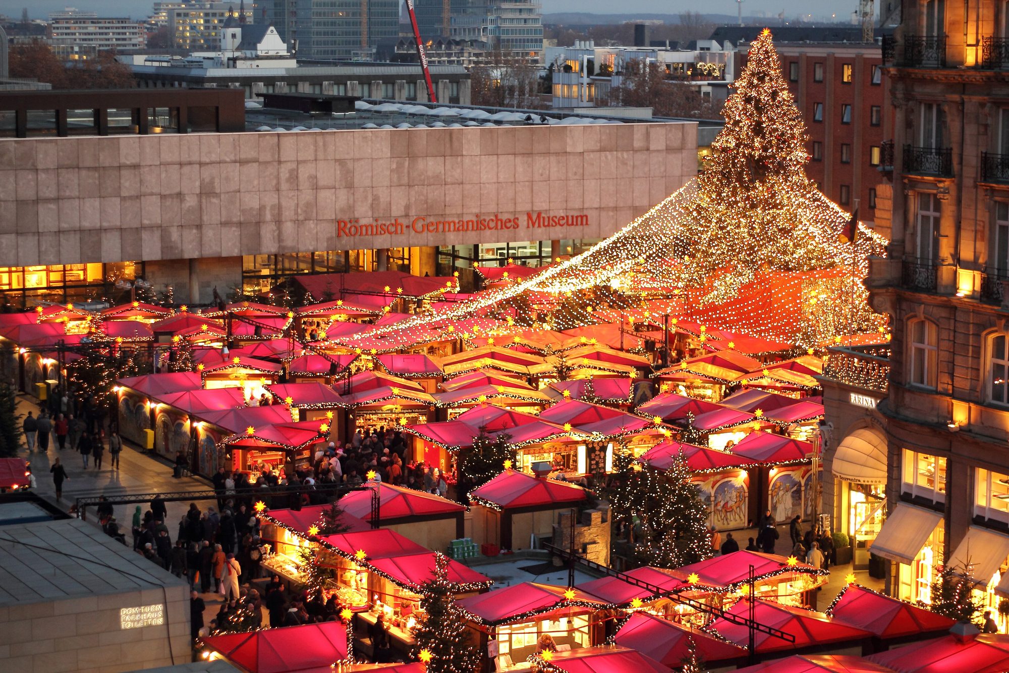 Rote Marktstände des Weihnachtsmarkts am Kölner Dom mit großem Lichterbaum in der Dämmerung