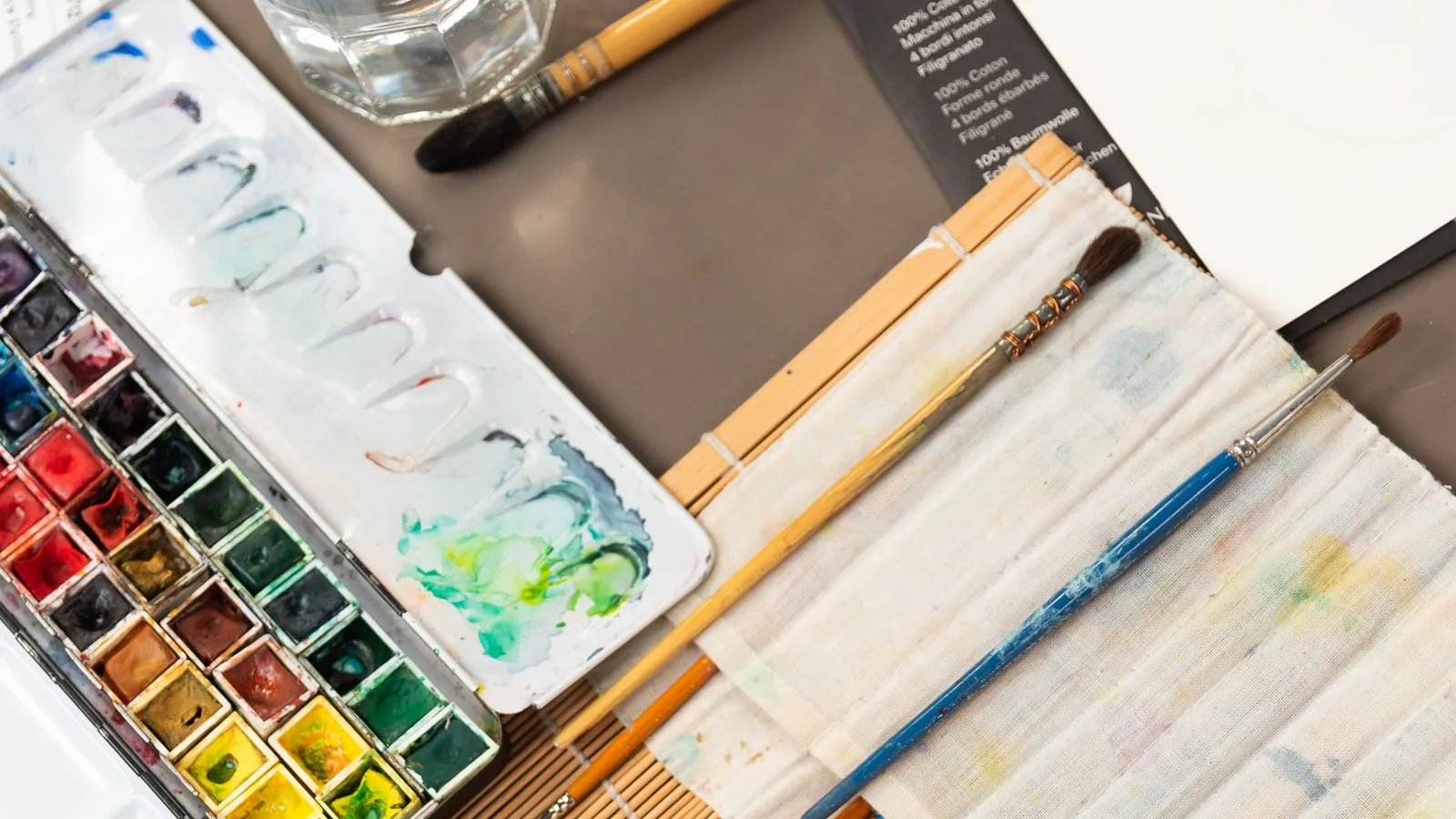 Aquarellkasten mit angemischten Farben, mehrere Pinsel in einer Stoffrolle und weißes Aquarellpapier auf einem ruhigen Arbeitstisch