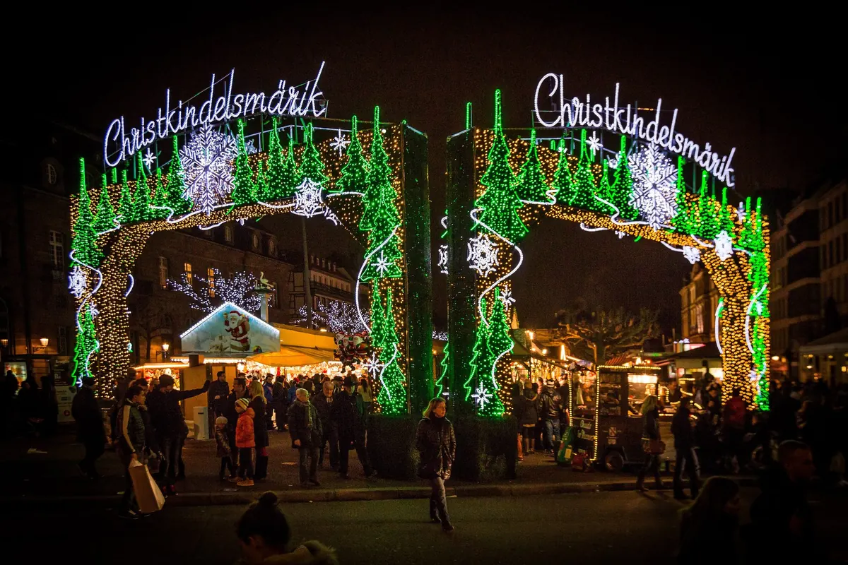 Beleuchteter Eingangsbogen des Christkindelsmärik in Straßburg am Abend, dahinter Marktstände